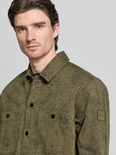 Männermodel trägt Regular Fit Hemdjacke aus reiner Baumwolle Modell 'LOCKY_2' von BOSS Orange in Oliv mit Allover-Muster, Label Patch, Nahaufnahme Oberkörper.
