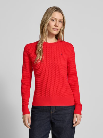 Frauenmodel trägt Slim Fit Pullover aus reiner Baumwolle von Tommy Hilfiger in Unifarbe Rot, Strickoptik mit Zopfmuster und Logo-Detail, Ganzkörperansicht von vorne.