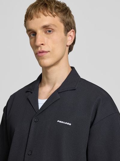 Männermodel trägt Relaxed Fit Freizeithemd mit Reverskragen Modell 'Troy' von Pegador in Schwarz mit Strukturmuster, Logo-Stitching, Knopfleiste, Nahaufnahme Oberkörper.