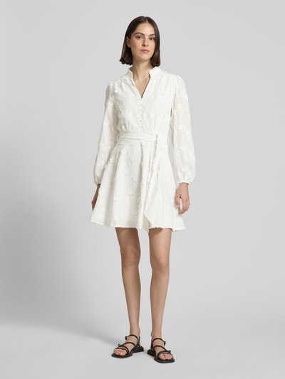 Frauenmodel trägt Minikleid mit Bindegürtel Modell 'Monnie' von Neo Noir in Weiß, Reine Baumwolle, Embroidery Design, Taillierter Schnitt, Serafino-Kragen, kurze Knopfleiste, Ganzkörperansicht von vorne.