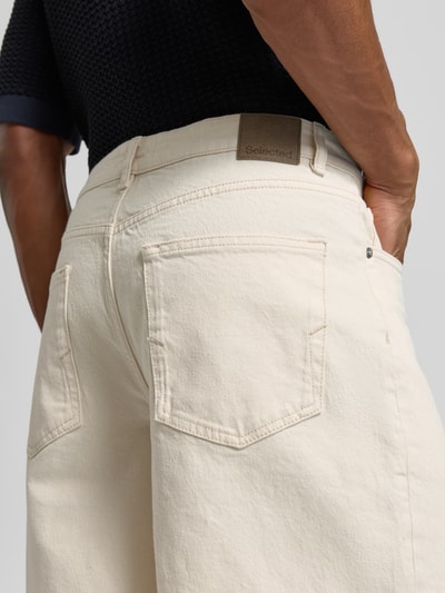 Männermodel trägt Relaxed Fit Jeansshorts aus Baumwoll-Mix Modell 'LOOSE ALEX SUMMER' von SELECTED HOMME in Offwhite, Twill-Struktur, Label Patch, 5-Pocket, Ganzkörperansicht Rückansicht.