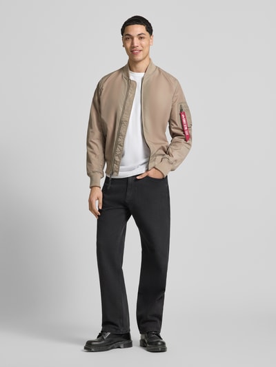 Męski model nosi Regular Fit Jacke mit Stehkragen od Alpha Industries, beżowa kurtka, czarne spodnie, jasnoszary t-shirt, gładka tekstura, widok całej sylwetki z przodu, kurtka z kieszenią na ramieniu, kieszeniami bocznymi, zamkiem błyskawicznym i elastycznymi mankietami, długość tyłu w rozmiarze M 66 cm, szerokość w rozmiarze M 48 cm (ramiona), 54 cm (klatka piersiowa), długość rękawa 68 cm.
