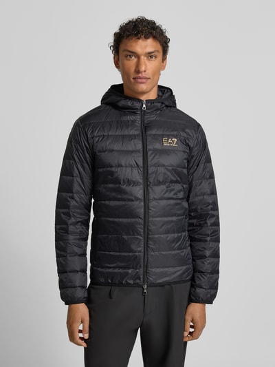 Männermodel trägt Daunenjacke mit Reißverschluss von EA7 Armani, schwarz, Regular Fit, Kapuze, Label-Print, Nahtreißverschlusstaschen, Ganzkörperansicht von vorne.