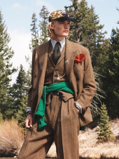 Männermodel trägt karierten braunen Anzug, gestreiftes Hemd mit grünem Pullover um die Taille und Krawatte, Ganzkörperansicht von vorne.