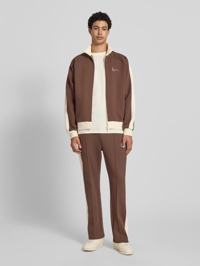Männermodel trägt Sweatjacke mit Stehkragen von KARL KANI in Braun mit Creme, Material-Mix, lockere Passform, Raglanärmel, Seitentaschen, Label-Stitching, Ganzkörperansicht von vorne.