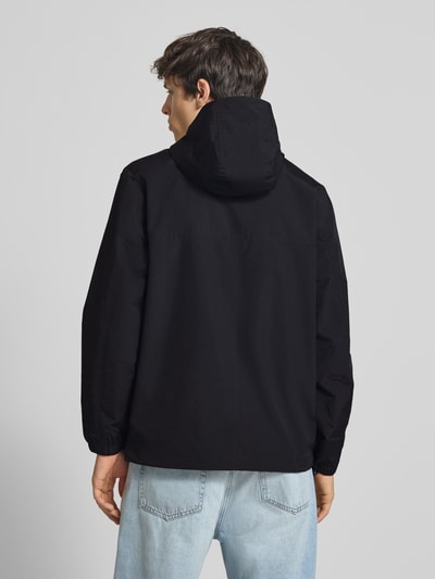 Männermodel trägt schwarzen Windbreaker mit Label-Stitching von Carhartt Work In Progress, Regular-Fit, Kapuze, Reißverschluss, Rückenlänge 73 cm, Schulterbreite 48 cm, Brustweite 55 cm, Ärmellänge 66 cm, Ganzkörperansicht Rückansicht.