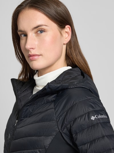 Kobiece modelka nosi Slim Fit Steppjacke mit Reißverschluss Modell 'Omni-Shield' von Columbia, czarny kolor, elastyczny materiał, krój slim fit, kaptur, kieszenie na zamek, elastyczne mankiety, długość tyłu w rozmiarze S: 65 cm, szerokość ramion: 44 cm, szerokość klatki piersiowej: 45 cm, długość rękawa: 60 cm, zbliżenie na górną część sylwetki.