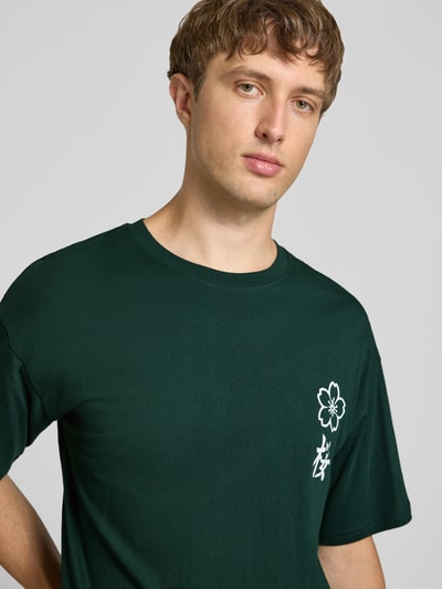 Männermodel trägt Relaxed Fit T-Shirt aus reiner Baumwolle Modell "DYLAN" von JACK & JONES in Dunkelgrün mit Print-Detail, Nahaufnahme Oberkörper.