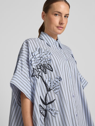 Frauenmodel trägt Oversized Kurzarmbluse aus reiner Baumwolle Modell 'JONKOE' von Essentiel Antwerp, hellblau mit Streifen und schwarzer Blumenstickerei, Nahaufnahme Oberkörper.