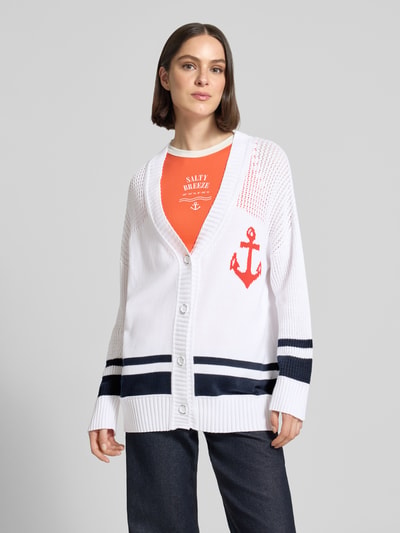 Frauenmodel trägt Cardigan mit überschnittenen Schultern und V-Ausschnitt von Marc Cain aus reiner Baumwolle, Loose-Fit, Knopfleiste, Rückenlänge 68 cm, Ärmellänge 48 cm, weiße Grundfarbe mit navyfarbenen und roten Details, Ganzkörperansicht von vorne.