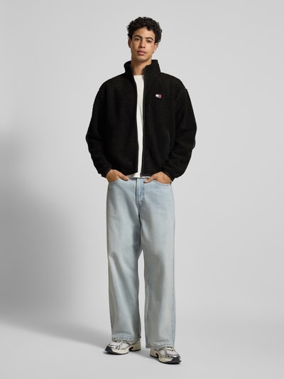 Männermodel trägt Relaxed Fit Fleecejacke in Teddyfleece-Optik von Tommy Jeans, Optik: Unifarben, Passform: Relaxed-Fit, Stehkragen, Reißverschluss, Label-Stitching, Logo-Detail, Langarm, elastischer Ärmel- und Rumpfabschluss, seitliche Eingrifftaschen, Materialverarbeitung: Webkunstfell, schwarze Jacke zu heller Jeans, Ganzkörperansicht von vorne.