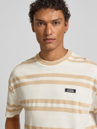 Męski model nosi T-shirt mit Label-Detail od Guess z bawełny, luźny krój, prążkowany okrągły dekolt, długość tyłu 71 cm, nahaufnahme górnej części ciała.