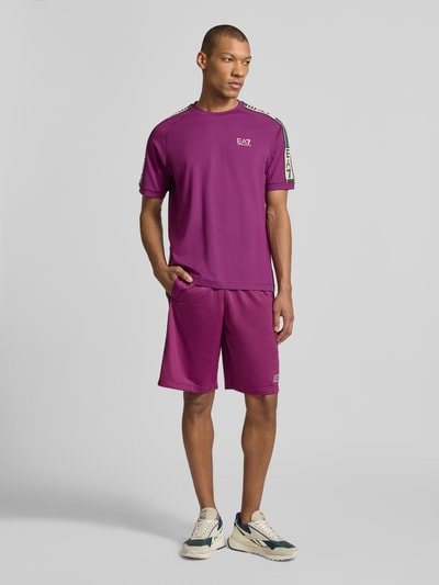 Męski model nosi T-shirt aus Viskose-Mix mit Logo von EA7 Emporio Armani w kolorze fioletowym, logo, krój regular fit, okrągły dekolt, długość tyłu w rozmiarze M: 70 cm, widok całej sylwetki z przodu.