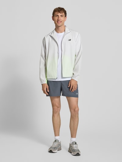 Männermodel trägt Regular Fit Jacke in Stehkragen Modell 'Fast Days' von New Balance mit Logo-Print, Reißverschluss, Eingrifftaschen, elastischen Ärmelabschlüssen und Tunnelzug im Bund, Farben Weiß und Hellgrün, glatte Textur, Ganzkörperansicht von vorne.