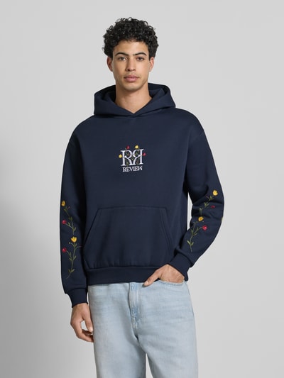 Männermodel trägt Regular Fit Hoodie mit Logo-Stitching von REVIEW, dunkelblau, florales Stitching, Känguru-Tasche, Ganzkörperansicht von vorne.