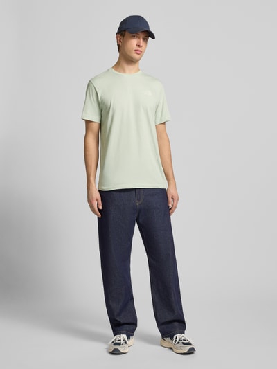 Męski model nosi T-shirt mit Label-Print od The North Face, mieszanka bawełny, krój regular fit, prążkowany okrągły dekolt, długość tyłu w rozmiarze M: 71 cm, jasne kolory, prosta forma, gładka faktura, Ganzkörperansicht von vorne.