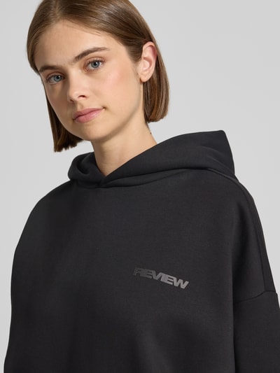Frauenmodel trägt Hoodie mit Logo-Print und Känguru-Tasche von REVIEW im Baumwoll-Mix, Loose Fit, Schwarz, Nahaufnahme Oberkörper.