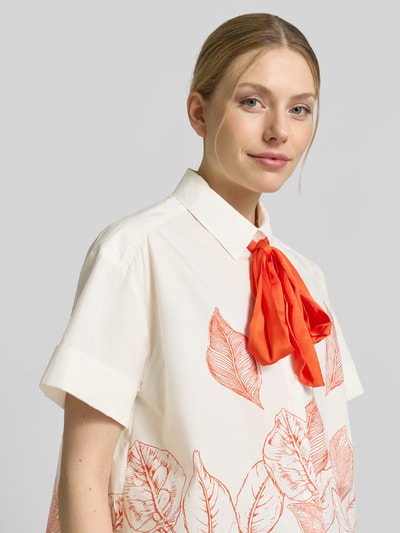 Frauenmodel trägt Regular Fit Schluppenbluse mit Motiv-Print Modell 'Saras' von MOS MOSH aus Reine Baumwolle, Nahaufnahme Oberkörper. Rückenlänge bei Größe S: 67 cm, Ärmellänge bei Größe S: 18 cm. Bluse mit cremefarbenem Grund, roten Blättermotiven und auffälliger roter Schleife.