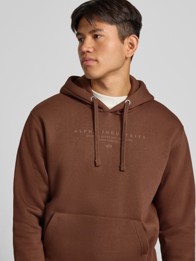 Männermodel trägt Relaxed Fit Hoodie aus Baumwoll-Mix mit Label-Detail von Alpha Industries in Braun, glatte Struktur, Nahaufnahme Oberkörper. Herren Hoodie von Alpha Industries, Känguru-Tasche, Kapuze mit Tunnelzug, elastische Abschlüsse, Länge bei Größe M 69 cm, Ärmellänge 62 cm.