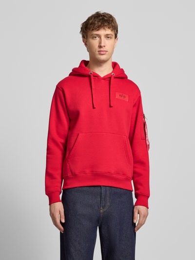 Männermodel trägt Hoodie mit Känguru-Tasche von Alpha Industries in Rot, Baumwoll-Mix, Regular-Fit, Label-Patch, Ganzkörperansicht von vorne.