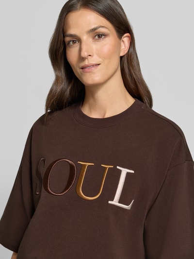 Frauenmodel trägt Cropped Boxy Fit Sweatshirt mit Label-Stitching von Smith and Soul, Rundhalsausschnitt, 1/2-Arm, Braun, Nahaufnahme Oberkörper.