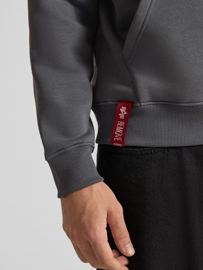 Männermodel trägt Hoodie mit Label-Details von Alpha Industries, Mieszanka bawełny, luźny krój, prążkowane wykończenia, kieszeń kangurka, detal z logo, kolor szary, Długość tyłu w rozmiarze M: 69 cm, Długość rękawów w rozmiarze M: 70 cm, zbliżenie na dolną część bluzy, Nahaufnahme Oberkörper.