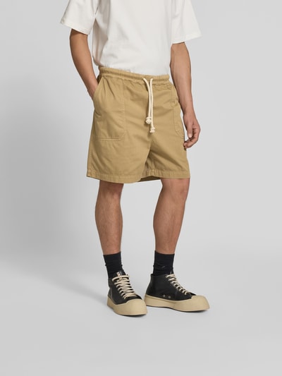 Männermodel trägt Bermudas aus reiner Baumwolle von Officine Générale, beige, lockere Passform, elastischer Bund, Eingrifftaschen, Tunnelzug, Ganzkörperansicht von vorne.