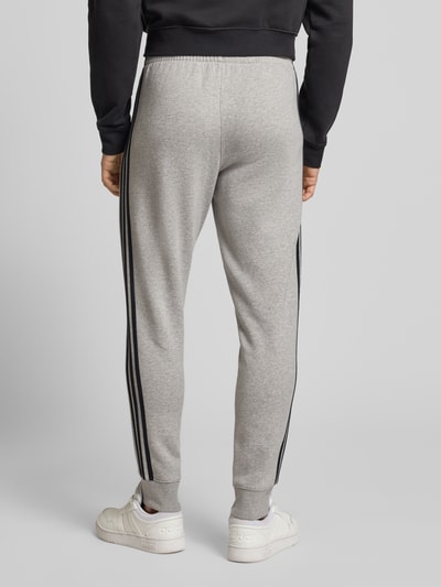 Männermodel trägt Sweatpants mit elastischem Bund von adidas Originals in lichtgrijs met zwarte strepen, katoenmix, los vallende fit, label-stitching, steekzakken, elastische tailleband, binnenbeenlengte maat M 75 cm, full body achteraanzicht.