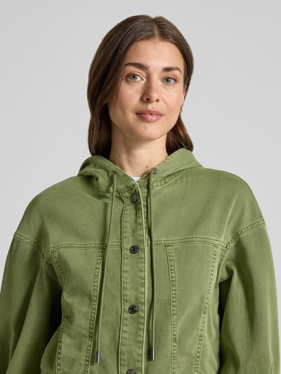 Modelka kobieca nosi Jeansjacke mit Kapuze und Eingrifftaschen od Marc Cain, z bawełnianej mieszanki ze stretchem, krój regular fit, zielony kolor, napy, kieszenie boczne, kaptur z troczkiem, szerokość w rozmiarze 36: 53 cm, długość tyłu: 51 cm, szerokość klatki piersiowej: 52 cm, długość rękawa: 54 cm, zbliżenie na górną część ciała.