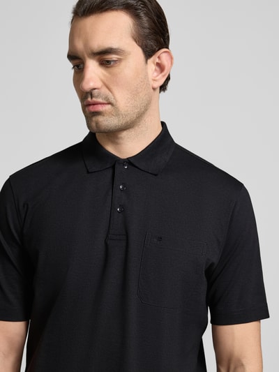 Mężczyzna nosi Regular Fit Poloshirt mit Brusttasche od Christian Berg Men, czarny materiał z bawełną, z kołnierzykiem, guziki, kieszeń na piersi, mieszanka materiałowa, krój regular fit, długość tyłu 68 cm w rozmiarze M, zbliżenie na górną część ciała.