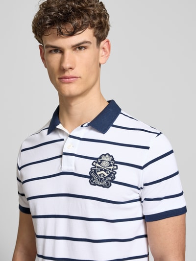 Männermodel trägt Slim Fit Poloshirt mit Label-Stitching und Knopfleiste von Superdry aus Reine Baumwolle, Nahaufnahme Oberkörper. Rückenlänge bei Größe M: 70 cm.