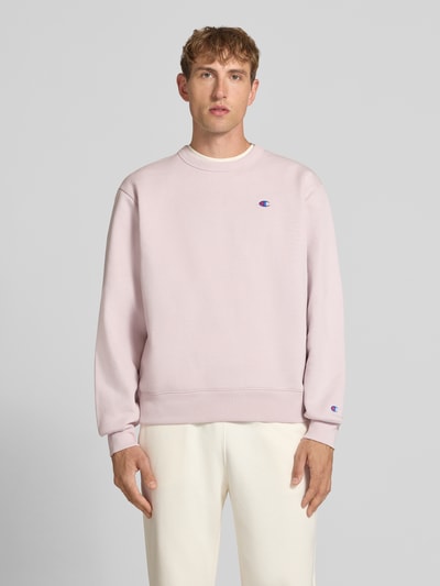 Männermodel trägt Sweatshirt mit Label-Details von Champion in blassem Rosa aus Baumwoll-Mix, lockere Passform, Rundhalsausschnitt, überschnittene Schultern, Ganzkörperansicht von vorne.