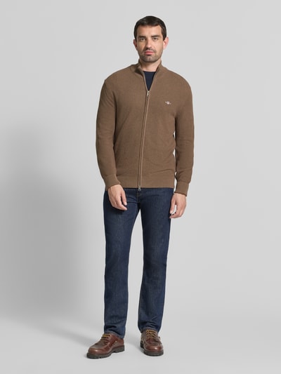 Männermodel trägt Regular Fit Strickjacke aus reiner Baumwolle von GANT in Braun, kombiniert mit Jeans, Ganzkörperansicht von vorne.