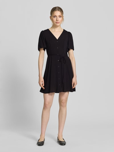 Frauenmodel trägt Leicht ausgestelltes Minikleid aus reiner Viskose Modell 'ALBA' von Vero Moda, Optik: Unifarben, Passform: Leicht ausgestellt, Mini-Länge, V-Ausschnitt, Taillengürtel, Knopfleiste, Ärmelform: Kurzarm, Material: Reine Viskose, glatte Struktur, schwarze Farbe, Ganzkörperansicht von vorne.