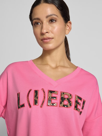 Vrouwenmodel draagt oversized sweatshirt van puur katoen Model 'L(I)EBE' van miss goodlife, V-hals, met pailletten, statementprint, ribboordjes. Nahaufnahme bovenlichaam.