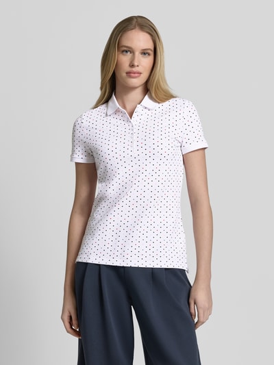 Modelka kobieca nosi Slim Fit Poloshirt mit Baumwoll-Anteil od Christian Berg Woman, bawełna z domieszką elastanu, krój slim fit, kołnierzyk polo, krótki rękaw, długość tyłu w rozmiarze S: 61 cm, biała tkanina w czarne kropki, proste linie, miękka tekstura, widok całej sylwetki z przodu.