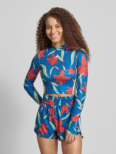 Frauenmodel trägt Slim Fit Sport-Langarmshirt mit Allover-Muster von adidas Sportswear, floral, Kontraststreifen, hochgeschlossen, AEROREADY, Materialverarbeitung: Interlock, kräftige Blautöne mit roten und grünen Akzenten, glatte Struktur, Optik: Allover-Muster, Ganzkörperansicht von vorne.