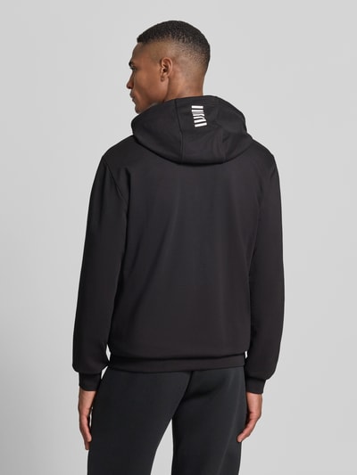 Męski model nosi Slim Fit Sweatjacke mit Kapuze od EA7 Emporio Armani, czarny kolor, elastyczna mieszanka bawełny, nadruk z logo, wpuszczane kieszenie, elastyczne wykończenia, długość tyłu 69 cm, długość rękawów 69 cm, krój slim fit, stójka, zamek błyskawiczny dwustronny, widok całej sylwetki od tyłu.
