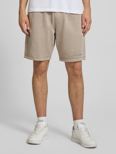 Männermodel trägt beigefarbene Regular Fit Sweatshorts mit Logo-Stitching von Pegador aus Baumwoll-Mix, elastischer Bund, Nahaufnahme Beine, Ganzkörperansicht von vorne.
