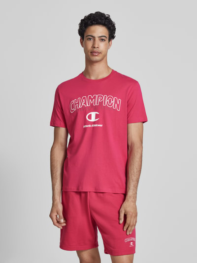Männermodel trägt T-Shirt mit Label-Print und Rundhalsausschnitt von CHAMPION in Rot, reine Baumwolle, Regular Fit, Nahaufnahme Oberkörper.