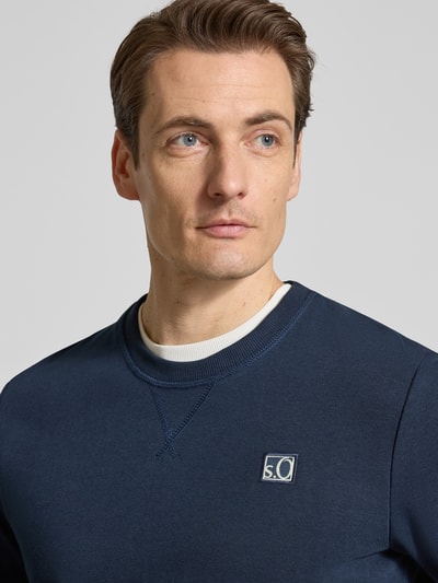 Męski model nosi Regular Fit Sweatshirt aus Baumwoll-Mix od s.Oliver RED LABEL, jednokolorowy, wyhaftowane logo, okrągły dekolt, mieszanka bawełny, dzianina dresowa, z długim rękawem, prążkowane wykończenie, kolor granatowy, tekstura gładka. Nahaufnahme górnej części ciała.