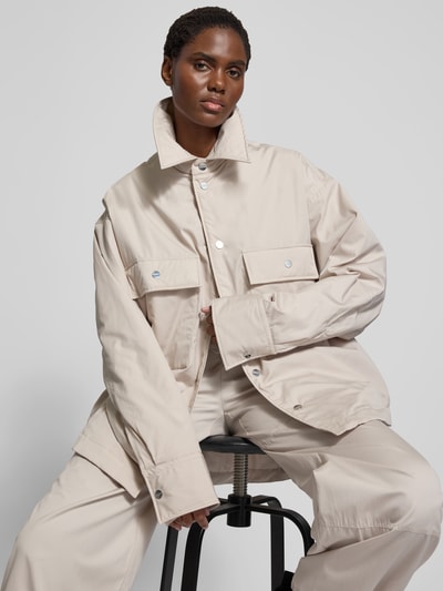 Frauenmodel trägt Jacke mit Hemdblusenkragen von black palms in Beige, Reine Baumwolle, Lockerer-Schnitt, Druckknopfleiste, Brusttaschen, Rückenlänge 79 cm bei Größe S, Nahaufnahme Oberkörper.