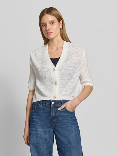 Modelka nosi kardigan mit V-Ausschnitt und 3/4-Arm von More & More, krój relaxed fit, listwa guzikowa, dekolt w serek, długość tyłu 48 cm, długość rękawów 24 cm, jasny kolor, faktura dzianiny, Ganzkörperansicht von vorne.