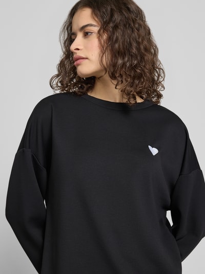 Frauenmodel trägt schwarzen Relaxed Fit Pullover mit weißer Herz-Stickerei Modell 'TALIA' von Pieces, unifarben, Nahaufnahme Oberkörper.