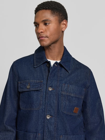 Männermodel trägt Regular Fit Jeansjacke aus reiner Baumwolle Modell 'WORK' von GANT, dunkelblau, Label-Patch, Nahaufnahme Oberkörper. Rückenlänge 71 cm, Schulterbreite 44 cm, Brustweite 51 cm, Ärmellänge 65 cm, aufgesetzte Eingriff- und Brusttaschen, Umlegekragen, durchgehende Knopfleiste.