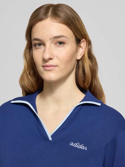 Frauenmodel trägt Poloshirt mit Label-Stitching von adidas Sportswear in Dunkelblau, reine Baumwolle, gerippte Ärmelabschlüsse, Nahaufnahme Oberkörper.
