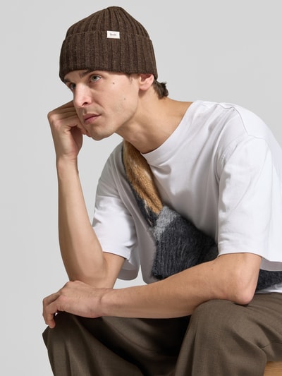 Männermodel trägt Beanie aus Woll-Mix von Forét in Braun, Ripp-Optik, breiter Umschlag, Label-Patch, Logo-Schriftzug, Nahaufnahme Oberkörper