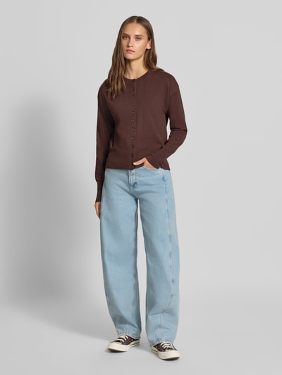 Kobiece modelka nosi brązowy kardigan rozpinany z mieszanki wiskozy Regular Fit Strickjacke aus Viskose-Mix model 'MIO' od ONLY, jasnoniebieskie jeansy, białe tenisówki, tło jasnoszare, prosty krój, tekstura dzianiny, widok całej sylwetki z przodu, damski kardigan ONLY.