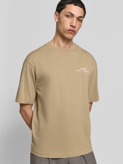 Männermodel trägt Relaxed Fit T-Shirt aus reiner Baumwolle von ANNARR in Beige mit Label-Print, Rundhalsausschnitt, kurzärmelig, Ganzkörperansicht von vorne. Rückenlänge bei Größe M: 67 cm.
