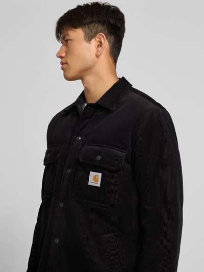 Männermodel trägt Regular fit Hemdjacke aus Cord Modell 'WHITSOME' von Carhartt Work In Progress, puur katoen, platte kraag, borstzak, zijzakken, labelpatch, zwarte kleur, structuur van corduroy, context: heren Hemdjacke, ruglengte maat M 77 cm, schouderbreedte 50 cm, mouwlengte 67 cm, nahaufnahme oberkörper.